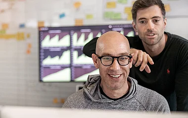 Zwei Männer stehen vor einem Bildschirm mit Diagrammen und diskutieren.