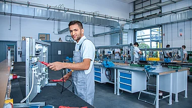 Techniker prüft elektrische Anlage mit Messgerät in einer Werkstatt.