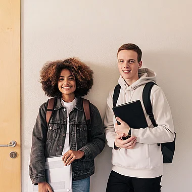 Zwei junge Erwachsene stehen lächelnd vor einer Tür mit Rucksäcken.