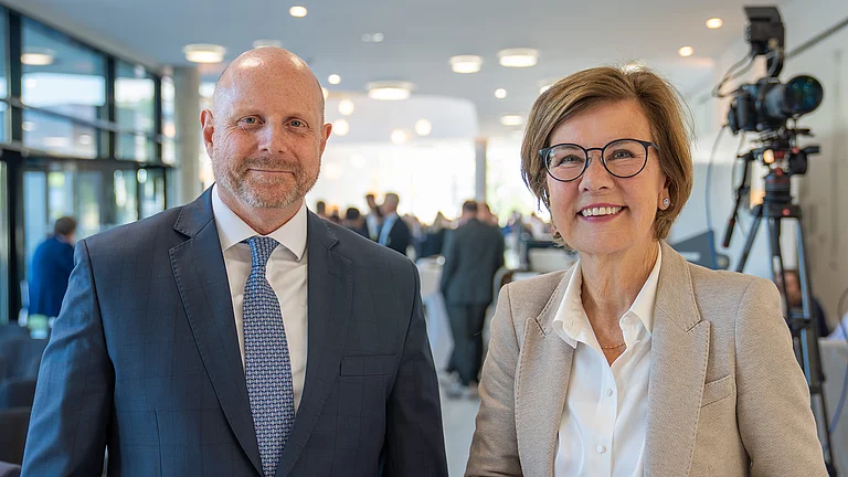 Ein Mann und eine Frau posieren lächelnd vor einer Konferenzkulisse.