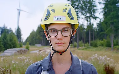 Person mit Schutzhelm und Brille steht vor einer Waldlichtung.