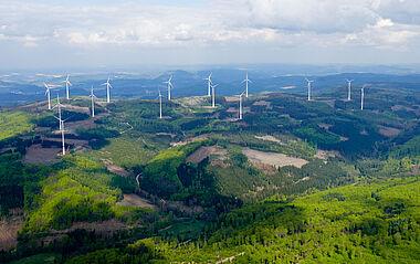 Windkraftanlagen in einer hügeligen Landschaft aus Wald und Feldern.