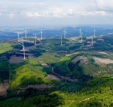Windkraftanlagen in einer hügeligen Landschaft aus Wald und Feldern.