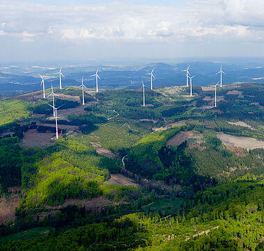Windkraftanlagen in einer hügeligen Landschaft aus Wald und Feldern.