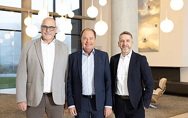 Drei Herren in Anzügen posieren lächelnd in einem modernen Büroambiente.