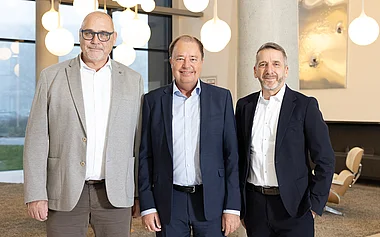 Drei Herren in Anzügen posieren lächelnd in einem modernen Büroambiente.