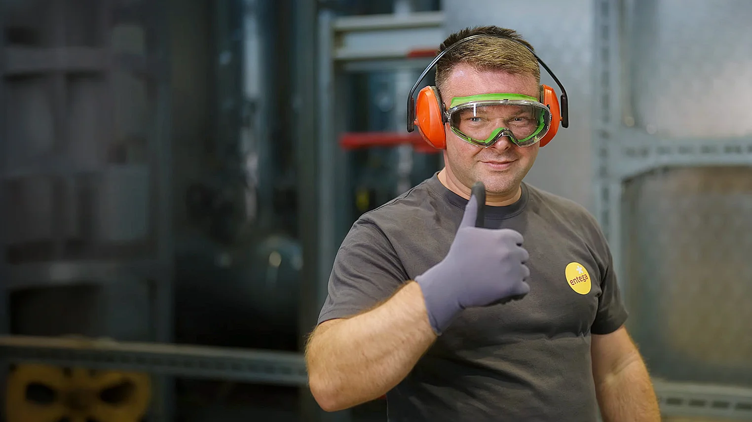 Techniker mit Schutzkleidung zeigt Daumen hoch vor Industrieanlagen.