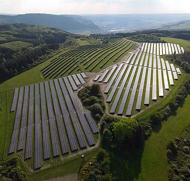 Solarmodule auf einer Hügelkuppe, umgeben von grüner Landschaft und bewaldeten Hängen.