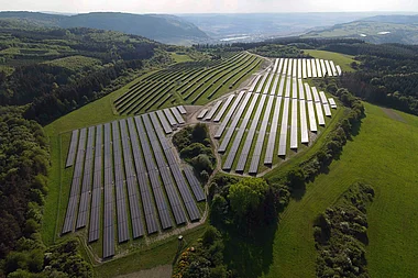 Solarmodule auf einer Hügelkuppe, umgeben von grüner Landschaft und bewaldeten Hängen.