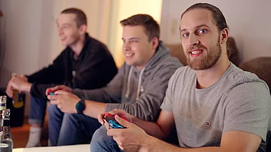 Drei junge Männer spielen lachend mit einem Videospiel-Controller im Wohnzimmer.