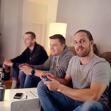 Drei junge Männer spielen lachend mit einem Videospiel-Controller im Wohnzimmer.