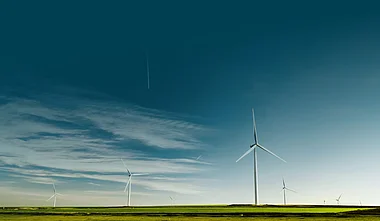 Windräder stehen auf einer grünen Hügelkuppe vor einem bewölkten Himmel.