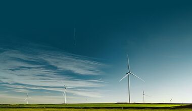 Windräder stehen auf einer grünen Hügelkuppe vor einem bewölkten Himmel.