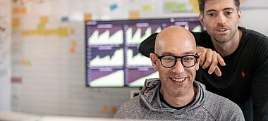 Zwei Männer stehen vor einem Bildschirm mit Diagrammen, vermutlich in einem Büro.