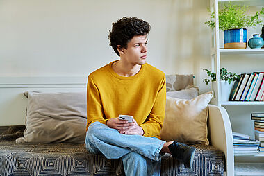 Ein Mann sitzt auf einem Sofa und hält ein Smartphone in der Hand.