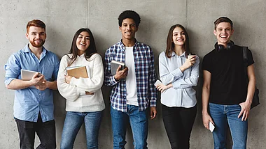 Vier junge Erwachsene stehen lächelnd in legerer Kleidung vor einer Betonwand.