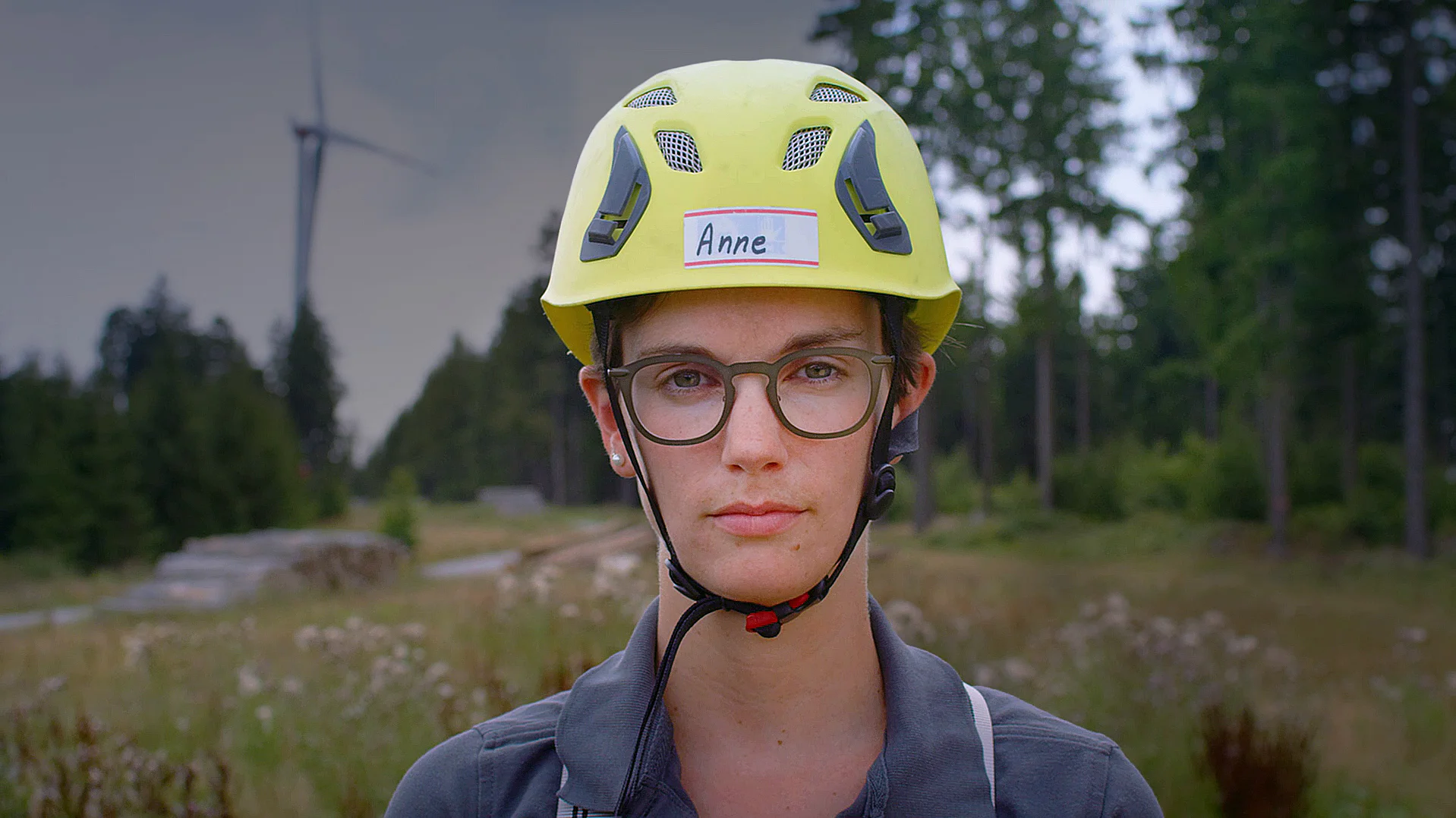 Eine Person mit Schutzhelm und Brille steht vor einer Waldlandschaft.