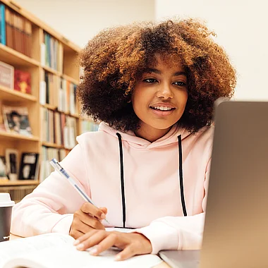 Eine junge Frau schreibt in ein Notizbuch, umgeben von Büchern.